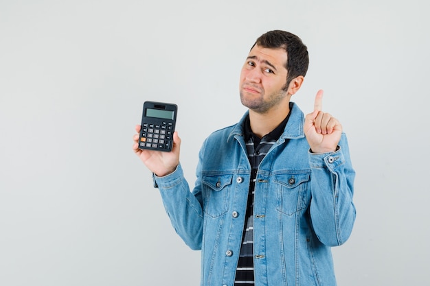 Hombre joven con calculadora, apuntando hacia arriba en camiseta, chaqueta y mirando decepcionado.