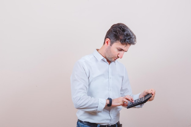 Hombre joven con calculadora en camisa blanca, jeans y mirando ocupado