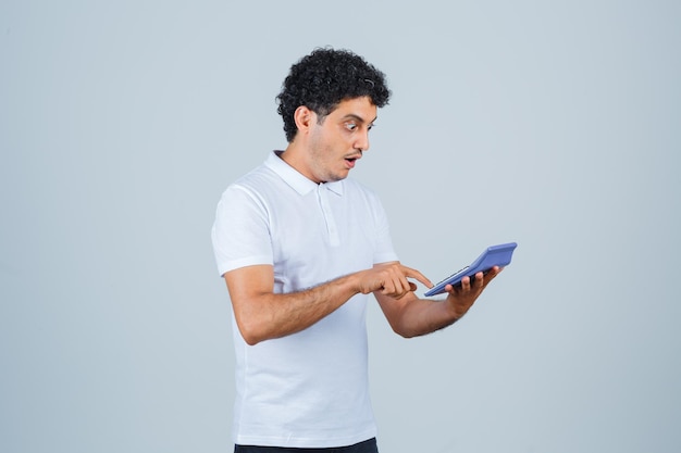 Hombre joven con calculadora en camiseta blanca y mirando sorprendido, vista frontal.
