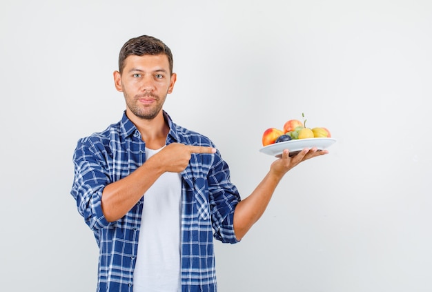 Foto gratuita hombre joven en camisa apuntando con el dedo a frutas en plato, vista frontal.