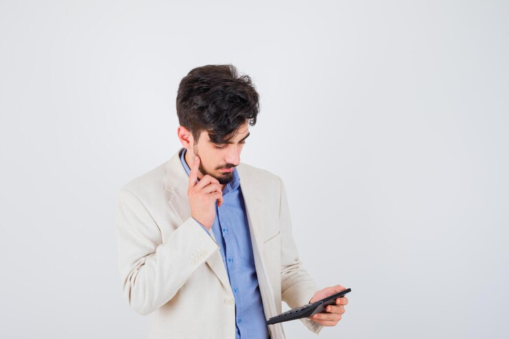 Foto gratuito hombre joven con camisa azul y chaqueta de traje blanco mirando calculadora y de pie en pose de pensamiento y mirando enfocado