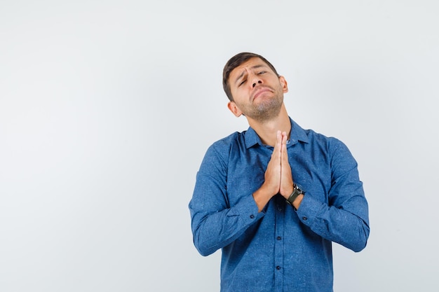 Hombre joven con camisa azul cogidos de la mano en gesto de oración y mirando indefenso, vista frontal.