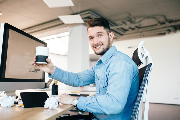 Hombre joven con camisa azul está sentado en su lugar de trabajo en la oficina. Viste camisa azul. Sostiene una taza y sonríe a la cámara.