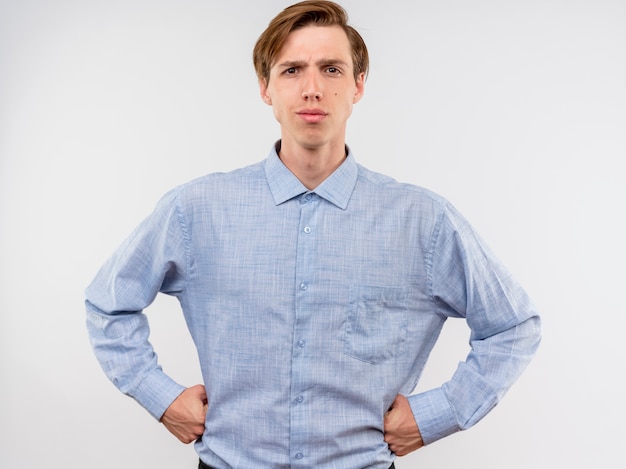 Hombre joven con camisa azul con expresión seria de confianza con los brazos en la cadera de pie sobre la pared blanca