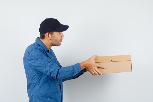 Hombre joven con camisa azul, gorra entregando caja de cartón y mirando alegre.