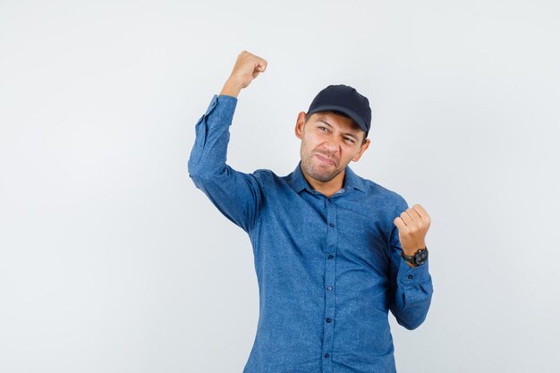 Hombre joven con camisa azul, gorra mostrando gesto de ganador y mirando feliz, vista frontal.