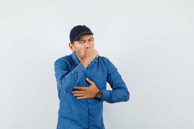 Hombre joven con camisa azul, gorra que sufre de dolor de garganta y tos, vista frontal.