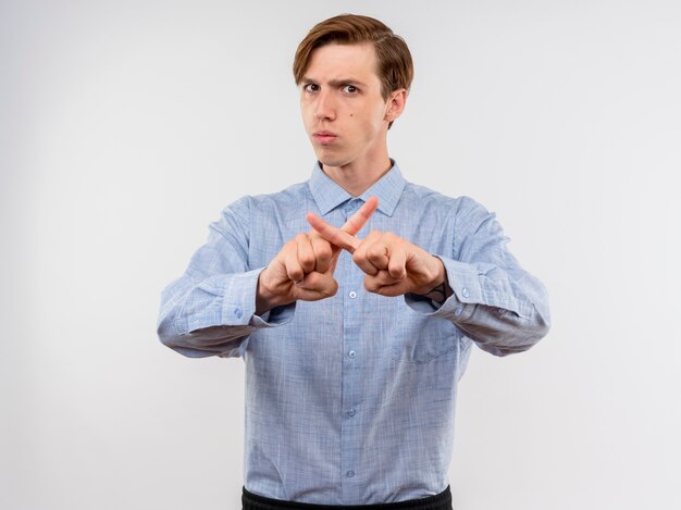 Hombre joven con camisa azul haciendo gesto de defensa cruzando los dedos índices con cara seria de pie sobre la pared blanca