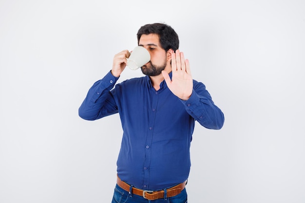 Hombre joven en camisa azul y jeans bebiendo una taza de agua y mostrando la señal de pare y mirando seria, vista frontal.