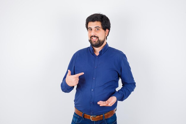 Hombre joven con camisa azul y jeans estirando la mano como sosteniendo algo y apuntando a sí mismo y mirando optimista, vista frontal.