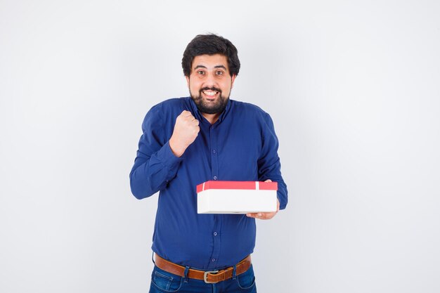Hombre joven con camisa azul y jeans sosteniendo una caja de regalo y apretando el puño y mirando feliz, vista frontal.