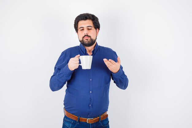 Hombre joven con camisa azul y jeans sosteniendo la taza y estirando la mano hacia él y mirando optimista, vista frontal.