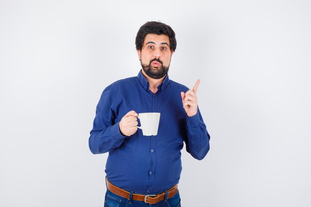 Hombre joven con camisa azul y jeans sosteniendo la taza y levantando el dedo índice en gesto eureka y mirando optimista, vista frontal.