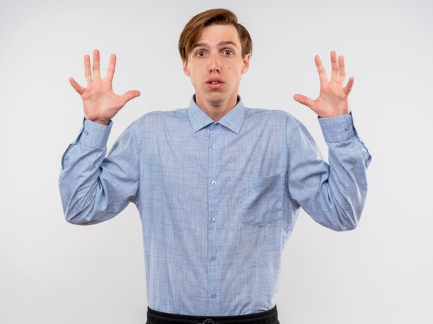 Hombre joven con camisa azul levantando las manos en la rendición de estar asustado de estar confundido de pie sobre la pared blanca