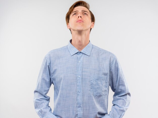 Hombre joven con camisa azul mirando hacia arriba con expresión seria de confianza de pie sobre la pared blanca