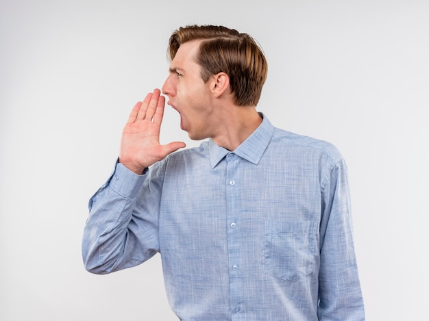 Hombre joven con camisa azul mirando a un lado gritando con la mano cerca de la boca de pie sobre la pared blanca