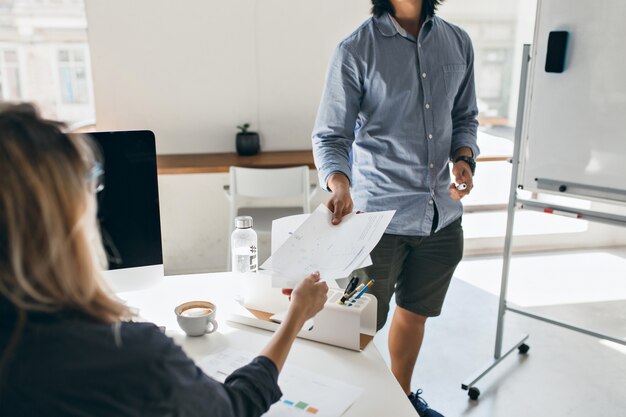 Hombre joven con camisa azul y pantalones cortos con documentos, caminando junto a la mesa de un colega. Retrato interior de una mujer rubia tomando café en la oficina y mirando el rotafolio.