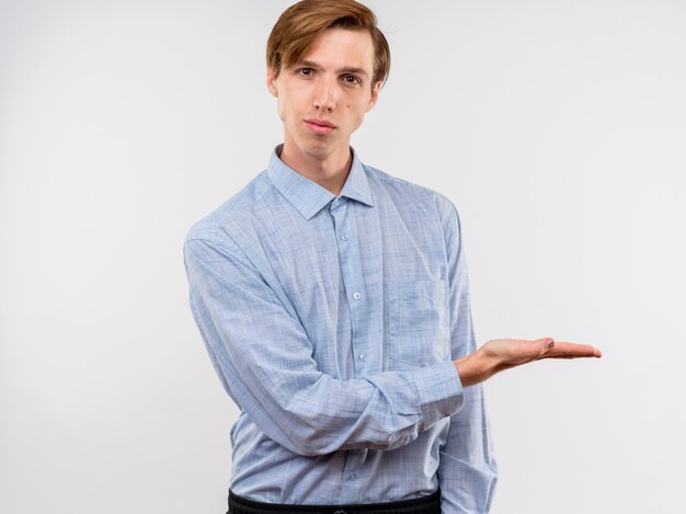Hombre joven con camisa azul que presenta algo con el brazo de su mano con expresión de confianza de pie sobre la pared blanca