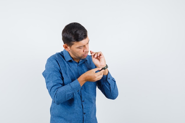 Hombre joven con camisa azul con reloj y mirando con cuidado, vista frontal.