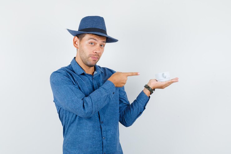 Foto gratuito hombre joven con camisa azul, sombrero apuntando a una taza de bebida y mirando alegre, vista frontal.
