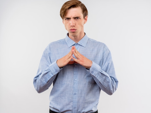 Hombre joven con camisa azul sosteniendo las palmas junto con cara seria esperando algo parado sobre la pared blanca