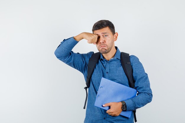 Hombre joven con camisa azul sosteniendo el portapapeles mientras se frota los ojos y mira triste, vista frontal.