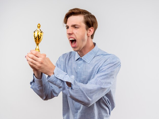 Foto gratuita hombre joven con camisa azul sosteniendo el trofeo mirándolo feliz y emocionado de pie sobre la pared blanca