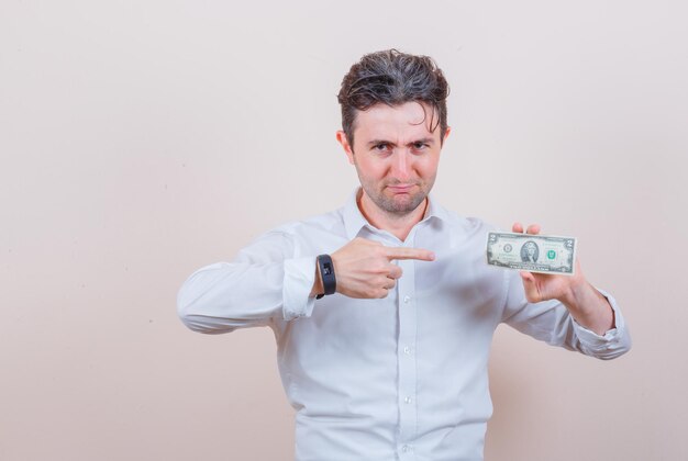 Hombre joven con camisa blanca apuntando al billete de un dólar y mirando positivo