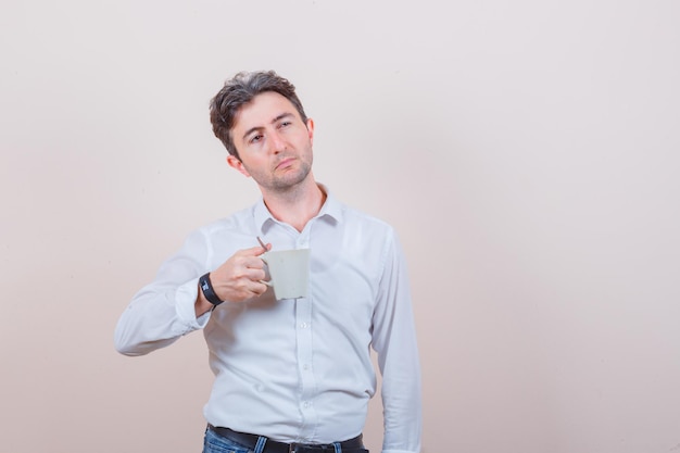 Hombre joven con camisa blanca, jeans sosteniendo una taza de bebida y mirando pensativo