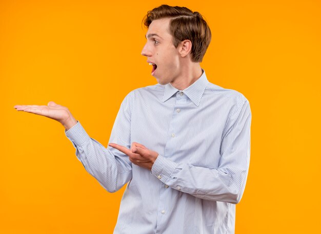 Hombre joven con camisa blanca mirando a un lado sorprendido y feliz presentando algo con el brazo de la mano apuntando con el dedo índice a algo parado sobre fondo naranja