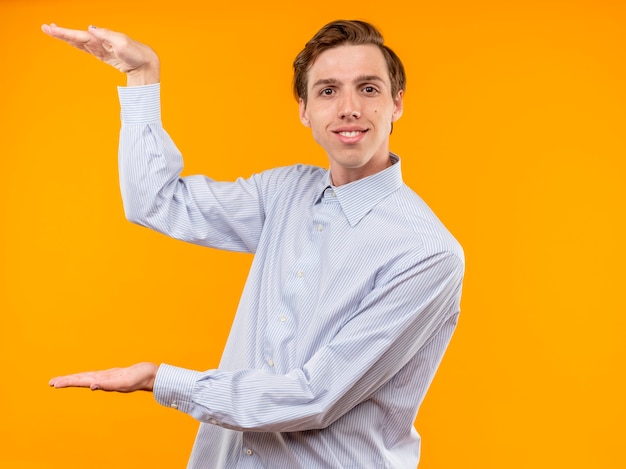 Hombre joven con camisa blanca mirando sonriendo con cara feliz mostrando gesto de tamaño con las manos, símbolo de medida de pie sobre la pared naranja