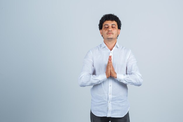 Hombre joven con camisa blanca mostrando gesto de namaste y mirando pacífica, vista frontal.