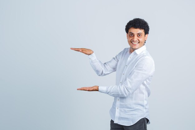 Hombre joven con camisa blanca, pantalones mostrando el signo de tamaño y mirando feliz, vista frontal.