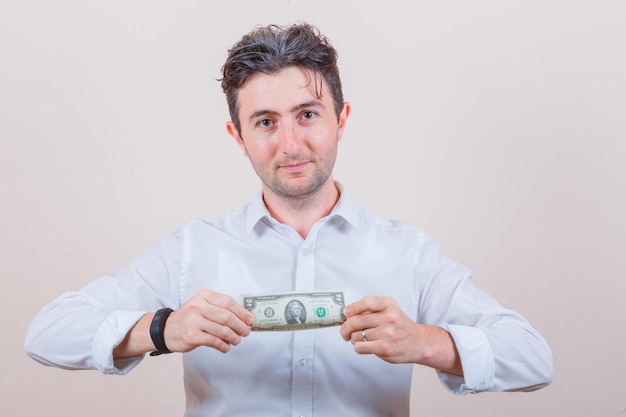 Hombre joven con camisa blanca sosteniendo billete de un dólar y mirando contento