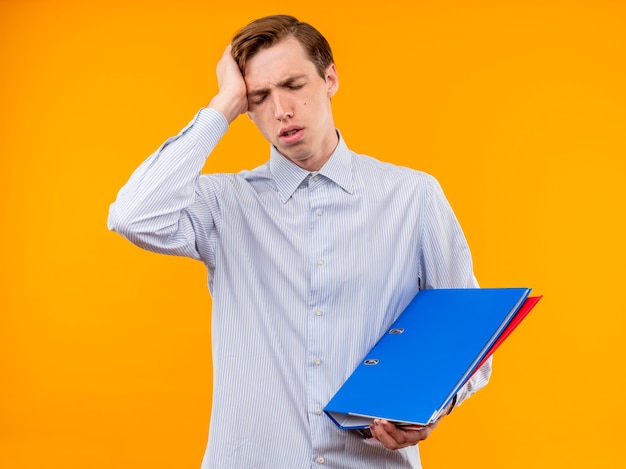 Foto gratuita hombre joven con camisa blanca sosteniendo carpetas mirando confundido y muy ansioso de pie sobre la pared naranja