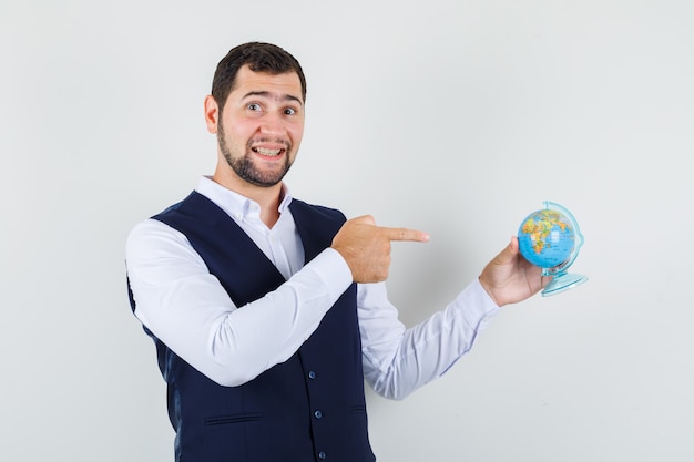 Hombre joven en camisa, chaleco apuntando al mundo y mirando alegre