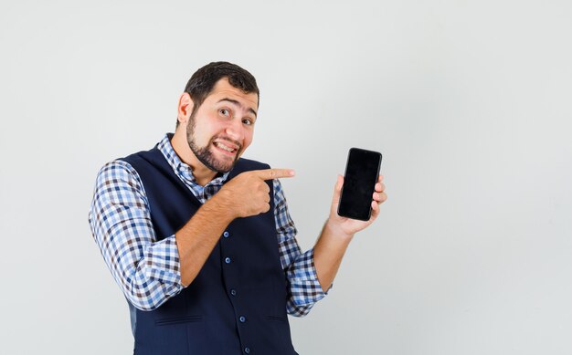 Hombre joven en camisa, chaleco apuntando al teléfono móvil y mirando alegre