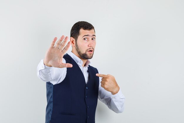 Hombre joven en camisa, chaleco que no muestra ningún gesto mientras se señala a sí mismo