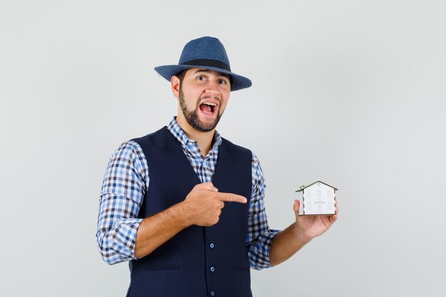 Hombre joven en camisa, chaleco, sombrero apuntando al modelo de la casa y mirando confiado, vista frontal.