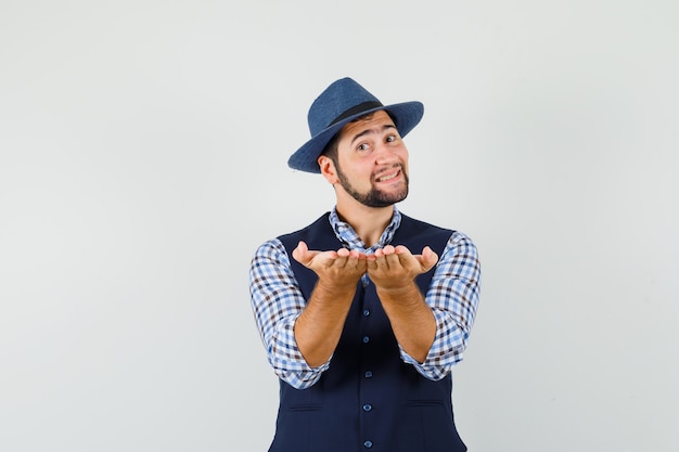 Hombre joven en camisa, chaleco, sombrero haciendo gesto de recepción o dar y mirando suave, vista frontal.