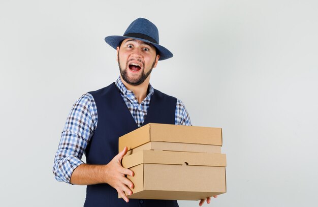 Hombre joven en camisa, chaleco, sombrero sosteniendo cajas de cartón y mirando alegre, vista frontal.