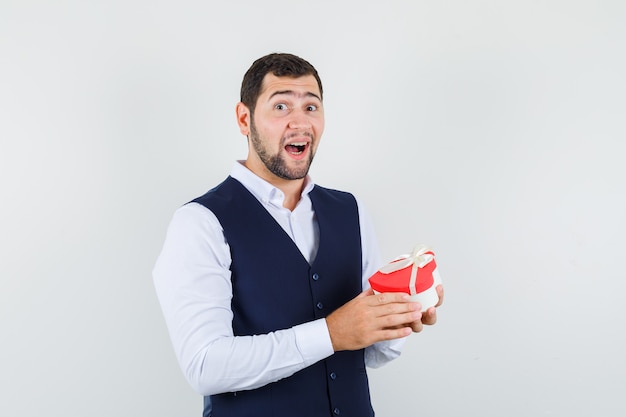 Hombre joven en camisa, chaleco sosteniendo la caja actual y mirando emocionado