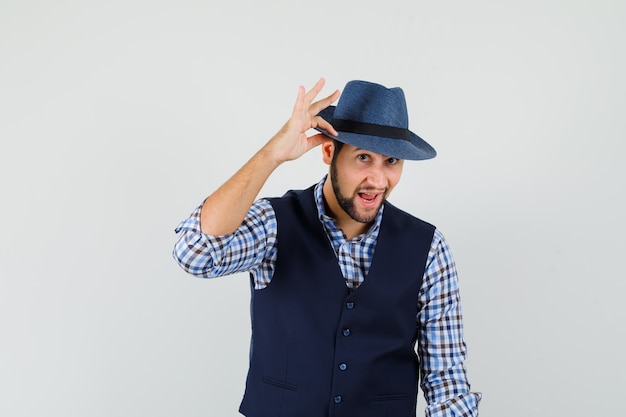 Foto gratuita hombre joven en camisa, chaleco tocando su sombrero con los dedos y luciendo elegante, vista frontal.