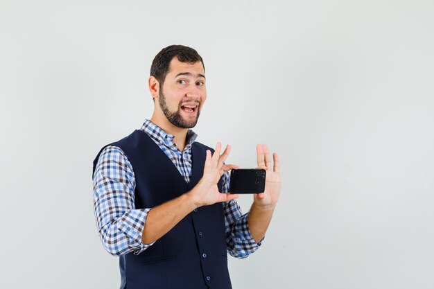 Hombre joven en camisa, chaleco tomando fotos en el teléfono móvil y mirando alegre, vista frontal.