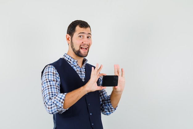 Hombre joven en camisa, chaleco tomando fotos en el teléfono móvil y mirando alegre, vista frontal.
