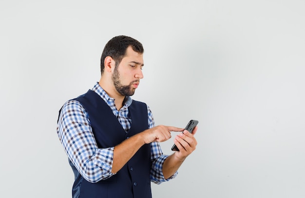 Hombre joven en camisa, chaleco usando teléfono móvil y mirando ocupado, vista frontal.