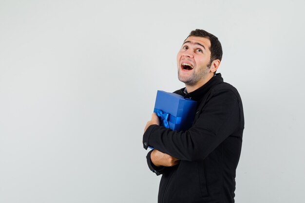 Hombre joven en camisa, chaqueta abrazando la caja actual y mirando feliz