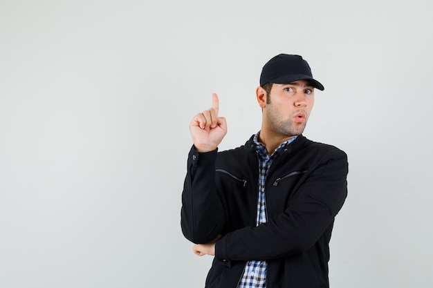 Hombre joven en camisa, chaqueta, gorra apuntando hacia arriba y mirando curioso