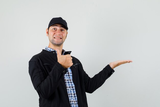 Hombre joven en camisa, chaqueta, gorra extendiendo la palma hacia un lado, mostrando el pulgar hacia arriba y mirando alegre