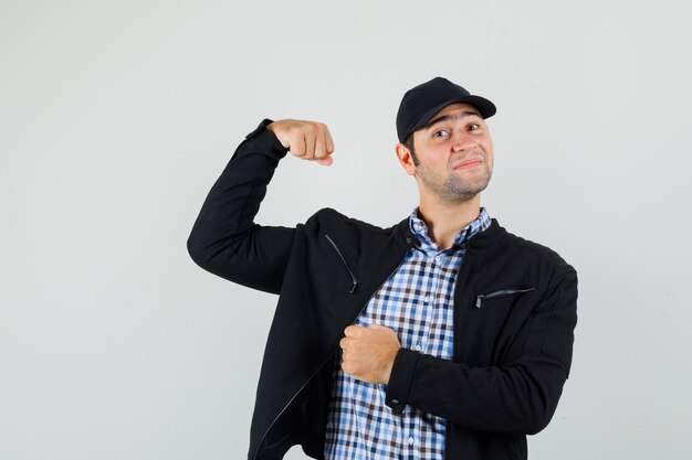 Hombre joven en camisa, chaqueta, gorra mostrando los músculos del brazo y mirando confiado
